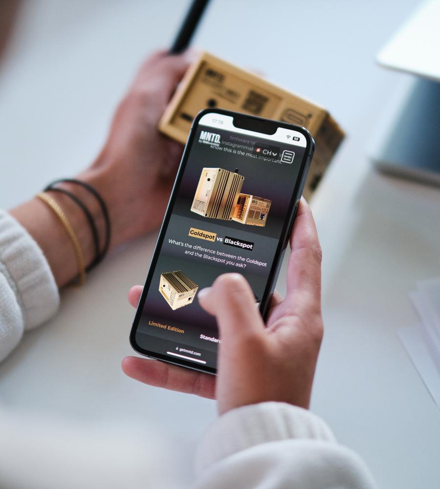 a human that holds a smartphone device in her hands and inside the screen we can see the online store of MNTD who sells crypto hardware and has been created by blokk studio. In the background we see a MNTD. gold hotspot